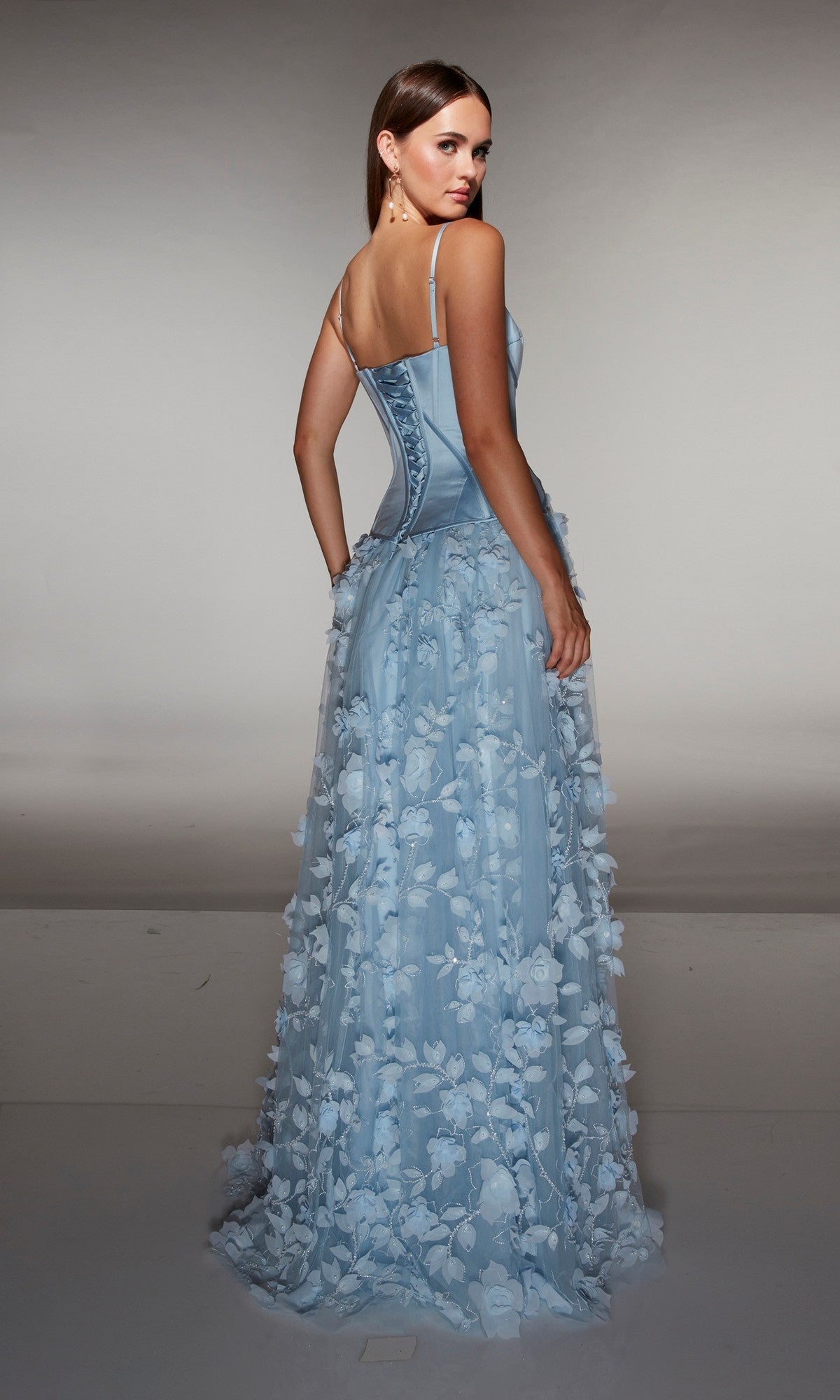A woman faces away in a studio, wearing the Alyce 62072 long formal dress—light blue with delicate thin straps, 3D floral-lace appliques, and a corset bodice.